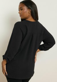 Maglietta nera a maniche lunghe con vestibilità rilassata, dotata di maniche arrotolabili e dettaglio con piega sul retro; texture morbida e tessuto leggero.