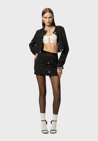 Giacca nera in tweed e mini gonna con bordi sfilacciati, top bianco corto e calze nere a strati. Presenta bottoni e dettagli in metallo argentato.