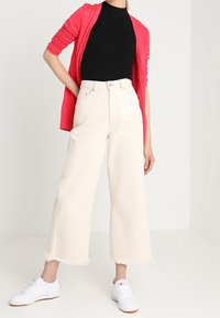 Weite cremefarbene Jeans mit rohem Saum, kombiniert mit einem figurbetonten schwarzen Rippoberteil und einem pinken Cardigan. Dazu weiße Sneakers.