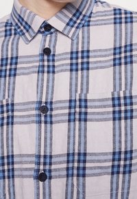 Lyhythihainen ruudullinen paita vaaleanbeigessä, jossa on sinisiä ja tummansinisempiä ruutukuvioita, napillinen kaulus sekä kaksi nappia edessä.