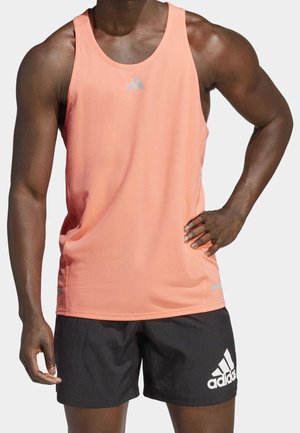 Personne musclée portant un débardeur de sport corail et un short noir Adidas, debout avec une main sur la hanche sur fond blanc.