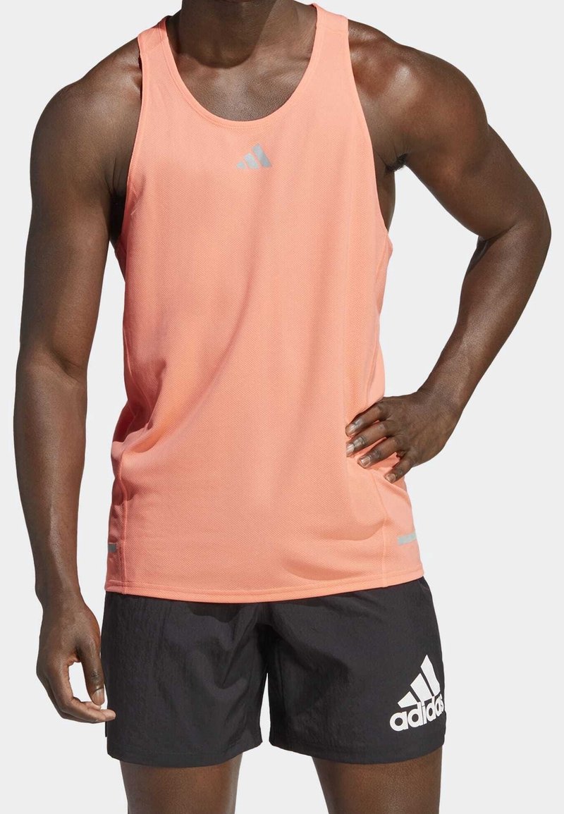 Personne musclée portant un débardeur de sport corail et un short noir Adidas, debout avec une main sur la hanche sur fond blanc.