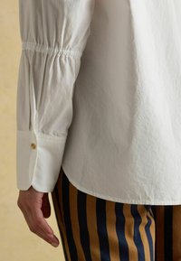 Weißes, strukturiertes Hemd mit gerafften Ärmeln und geknöpften Manschetten, kombiniert mit navy- und senfgelben gestreiften Hosen. Seitenansicht der Details des Kleidungsstücks.