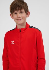 Red zip-up jacket with textured fabric. Features black patterned accents on shoulders and a white logo on the left chest.