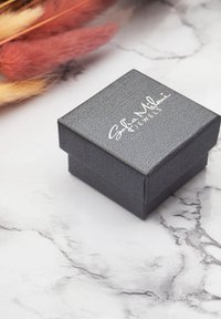 Schwarze, strukturierte Schmuckschatulle mit einem Klappdeckel und weißem, geprägtem Logo. Auf einer hellen Marmoroberfläche platziert, mit rosa getrockneten Blumen in der Nähe.