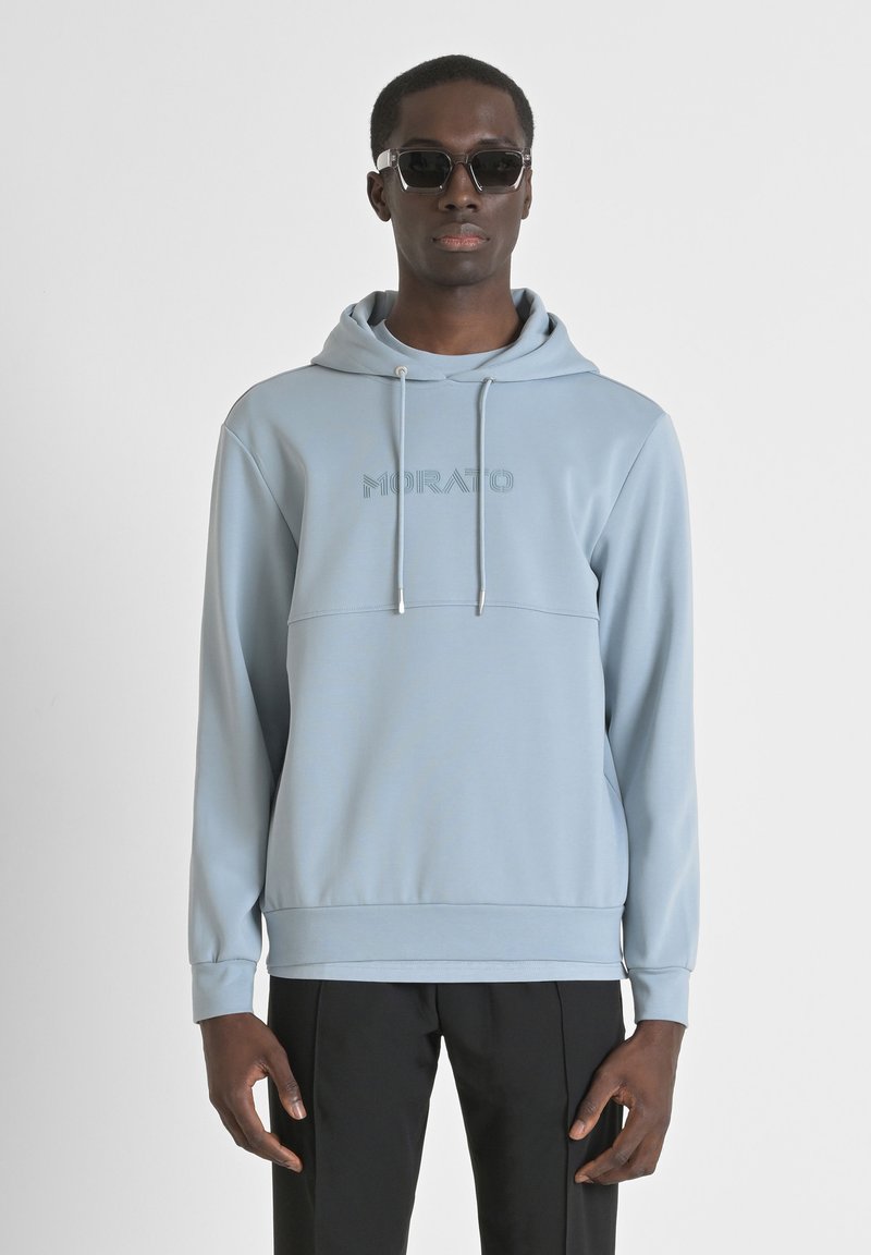 Felpa con cappuccio azzurro chiaro con logo "MORATO" tono su tono, con cordino, texture morbida e polsini a costine, abbinata a pantaloni neri e occhiali da sole.