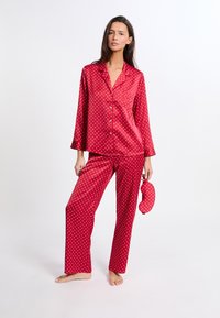 Conjunto de pijama de satén rojo con lunares blancos; camisa de manga larga, pantalones a juego y una máscara para dormir. Textura suave, corte relajado.