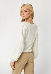 Suéter blanco de punto con textura acanalada, mangas abullonadas y un escote redondo. Combinado con pantalones ajustados color beige, mostrando un estilo casual.