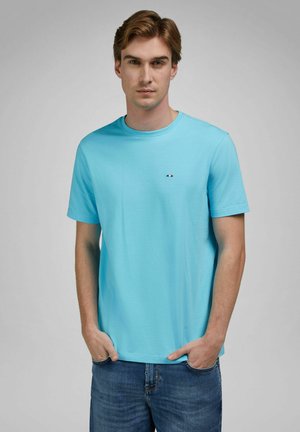 Jonge man met lichtbruin haar, gekleed in een lichtblauw T-shirt met korte mouwen en een blauwe spijkerbroek, handen in de zakken, neutrale grijze achtergrond.
