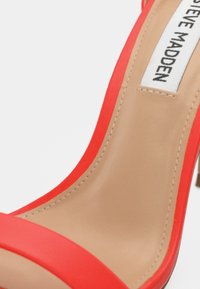 Κόκκινο σανδάλι με ψηλό τακούνι, λείο και γυαλιστερό φινίρισμα, με στενό λουράκι και μπεζ πάτο. Η ετικέτα είναι ορατή στον πάτο.