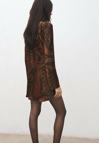 Brązowa sukienka z długim rękawem z czarnymi wzorami paisley, gładka w dotyku i o luźnym kroju, noszona z przezroczystymi czarnymi rajstopami.