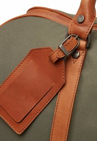 Sac vert olive avec une sangle en cuir lisse et une étiquette rectangulaire en cuir. Des accessoires en métal fixent la sangle au sac.