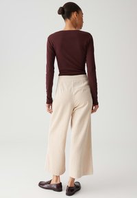 Vrouw met een knot in haar haar, gekleed in een strakke bordeauxrode top met lange mouwen, crèmekleurige wijde broek en donkerbruine instappers, staand tegen een effen achtergrond.