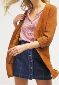 Veste en suède marron sur une chemise à motifs roses et une jupe en denim bleu avec des boutons. Présente une coupe décontractée et des manches retroussées.