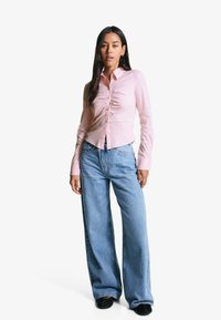 Camicia a maniche lunghe rosa chiaro con bottoni e dettagli arricciati, abbinata a jeans blu in denim a gamba larga e scarpe nere. Design semplice e pulito.