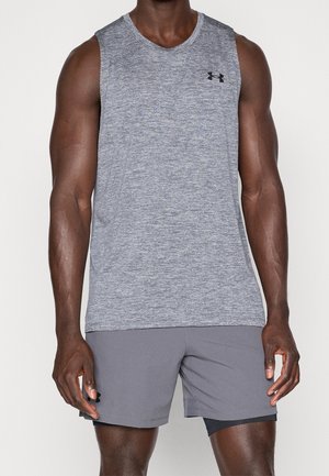 Personne musclée portant un haut de sport gris sans manches et un short gris assorti avec une couche inférieure noire, debout sur un fond blanc.