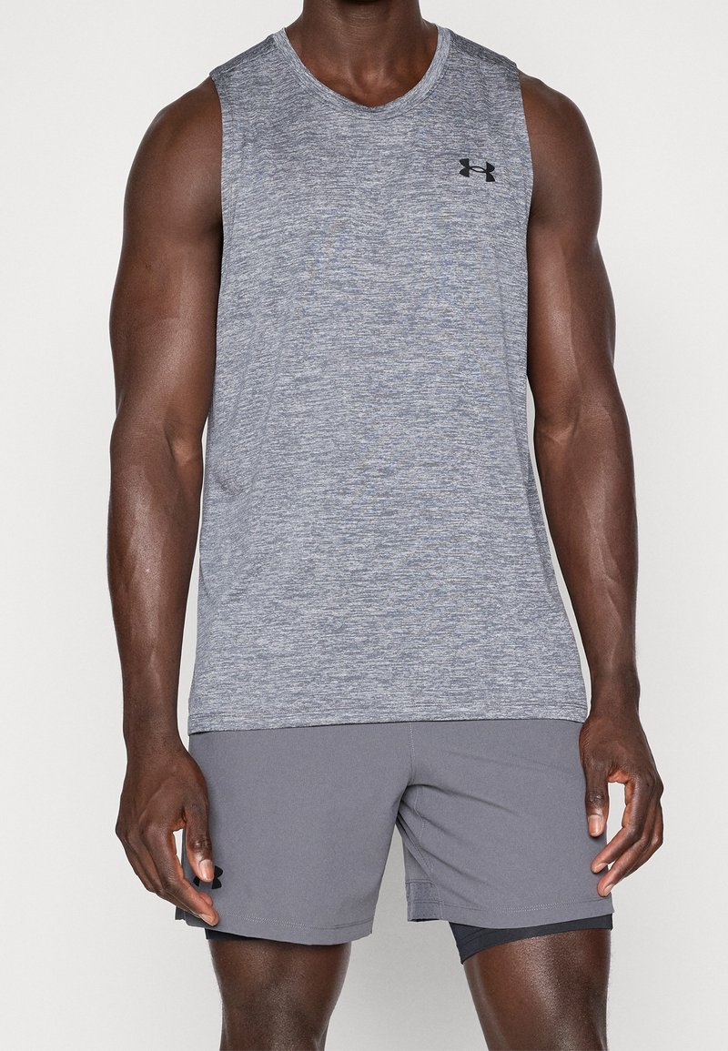 Personne musclée portant un haut de sport gris sans manches et un short gris assorti avec une couche inférieure noire, debout sur un fond blanc.