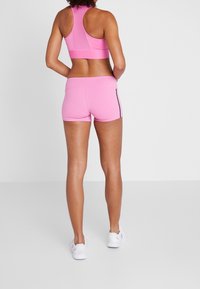 Růžová sportovní podprsenka a šortky s přiléhavým střihem, hladkou texturou a černými bočními akcenty. Bílá obuv doplňuje outfit.