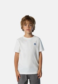 Weißes Baumwoll-T-Shirt mit Rundhalsausschnitt, kurzen Ärmeln und einem blauen Logo-Patch auf der Brust. Graue Shorts sind teilweise sichtbar.