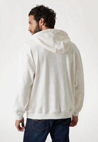 Hvid hættebluse med tekstureret mønster, der har en ensfarvet hætte og manchetter. Båret med mørkeblue jeans, hvor den ene hånd er placeret på hoften.