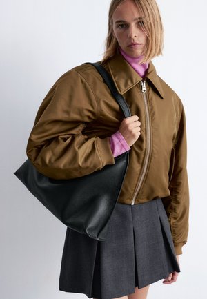 Brown bomber jacket with a zip closure, paired with a light pink turtleneck, gray pleated skirt, and a large black leather tote bag.
