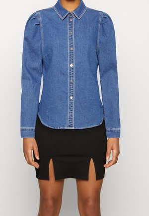 Kobieta w niebieskiej dżinsowej koszuli na guziki z bufiastymi ramionami i czarnej mini spódnicy z rozcięciami z przodu.