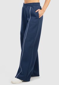 Marineblaue Sweatpants mit weitem Bein, elastischem Bund, seitlichen Taschen und weißen Paspeln entlang der Nähte. Aus weichem Stoff gefertigt.
