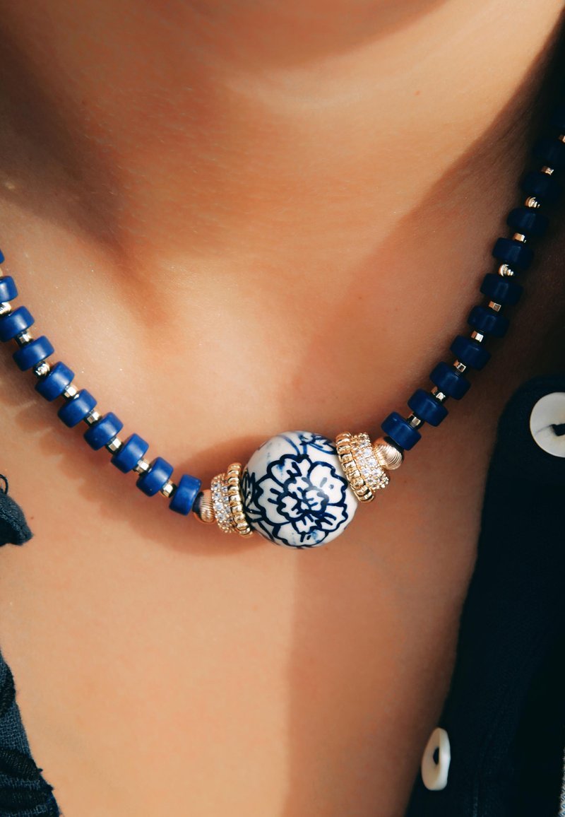 Collier en perles composé de perles rectangulaires bleues et d'une perle ronde centrale ornée d'un design floral bleu et blanc, accentué de cercles dorés.