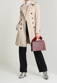 Béžový trenčkot s dvouřadými knoflíky, černé kalhoty a kotníkové boty ze vzoru hadí kůže. Drží bordó kabelku s červenými kožešinovými doplňky.