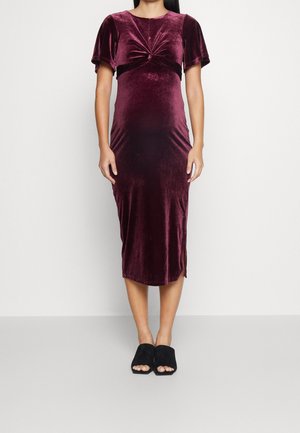 Kobieta ubrana w aksamitną bordową sukienkę z krótkim rękawem, sięgającą kolan, z detalem skręconego węzła na wysokości piersi oraz czarne sandały typu mule z odsłoniętymi palcami.