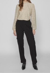Donna che indossa un top a maniche lunghe in pizzo beige, pantaloni cargo neri e stivali con tacco neri alla caviglia, in piedi davanti a uno sfondo neutro.