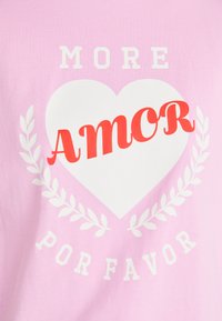 Růžové bavlněné tričko s bílým srdcem a červeným textem "AMOR", lemované bílými vavřínovými listy a frází "MORE POR FAVOR" nad ním.