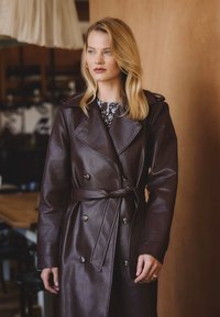 Brązowy skórzany płaszcz trench z podwójnymi guzikami, szerokimi klapami i paskiem w talii. Gładka tekstura z lekko błyszczącym wykończeniem.