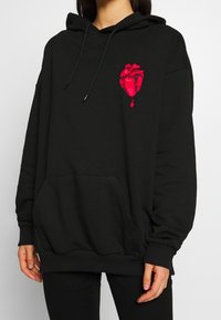 Persona que lleva una sudadera negra de gran tamaño con un gráfico de un corazón anatómico rojo y una gota roja en el lado izquierdo del pecho.