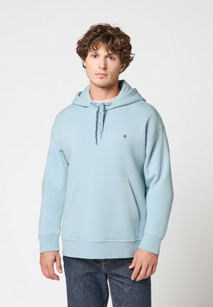 Jonge man met krullend haar die een lichtblauwe hoodie met voorzak en patroonkoorden draagt, staand tegen een effen witte achtergrond.