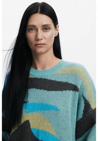 Pullover lavorato a maglia accogliente con base turchese e motivi astratti in nero, marrone e giallo. Collo rotondo e vestibilità comoda.