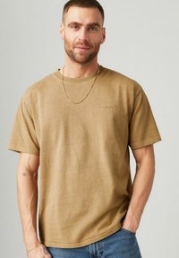 Beige Baumwoll-T-Shirt mit lockerer Passform, kurzen Ärmeln und rundem Halsausschnitt. Mit dezentem Stickdesign und glatter Textur. Kombiniert mit blauen Jeans.