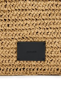 Borsa intrecciata in paglia con una finitura naturale e texture. Presenta un'etichetta rettangolare in morbida pelle nera con testo impresso, colori e materiali a contrasto.