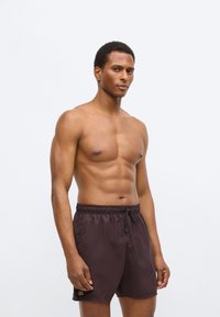 Shorts de bain marron avec une ceinture élastique et un cordon de serrage, arborant un petit logo vert sur l'ourlet gauche. Matière lisse et légère.