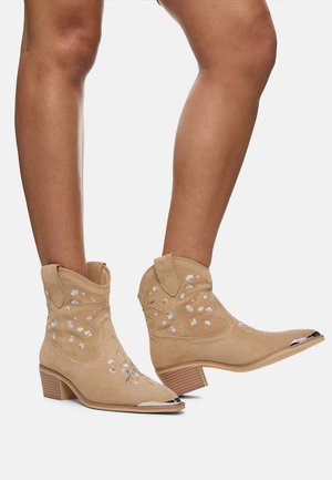Botas de vaquero de ante marrón con bordados florales, punteras afiladas y acentos plateados. Tacones cortos en bloque y detalles de costura decorativa.