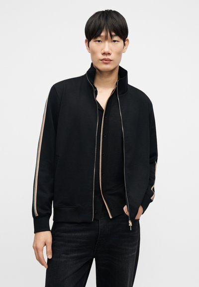 Μαύρο ζακέτα τζάκετ με φερμουάρ και ψηλό γιακά, με πολύχρωμες ρίγες κατά μήκος των μανικιών, συνδυασμένο με μαύρο πουκάμισο με κουμπιά και σκουρόχρωμο τζιν.