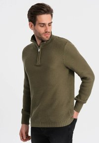 Olijfgroene gebreide pullover met een hoge kraag en een ritssluiting. Textuurpatroon en geribbelde manchetten voegen detail toe. Draagt met een donkere spijkerbroek.