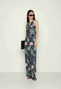 Macacão floral em azul marinho com um design de decote halter, pernas largas, e uma mistura de flores rosas e verdes. Acompanhado de uma clutch preta.