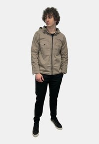 Giacca khaki con cappuccio e zip frontale, due tasche al petto e pantaloni neri. Tessuto testurizzato, design casual e stile a strati. Scarpe nere.