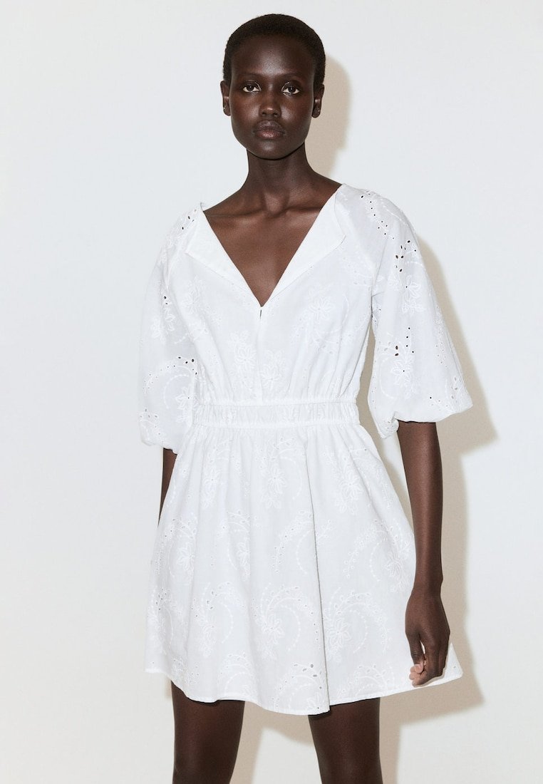 Femme à la peau foncée portant une robe courte blanche brodée avec des manches bouffantes, debout devant un fond clair uni.