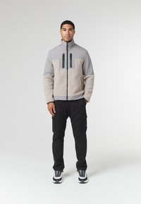 Bunda s fleece podšívkou, s šedou hornou časťou a béžovou sherpa dolnou časťou, s čiernymi zipsovým vreckami, nosená s čiernymi cargo nohavicami a teniskami.