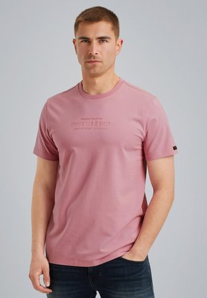 T-shirt kratkih rukava, okruglog izreza u svijetlo ružičastoj boji od pamuka s urezanim tekstom brenda na prsima, u kombinaciji s tamnim trapericama.