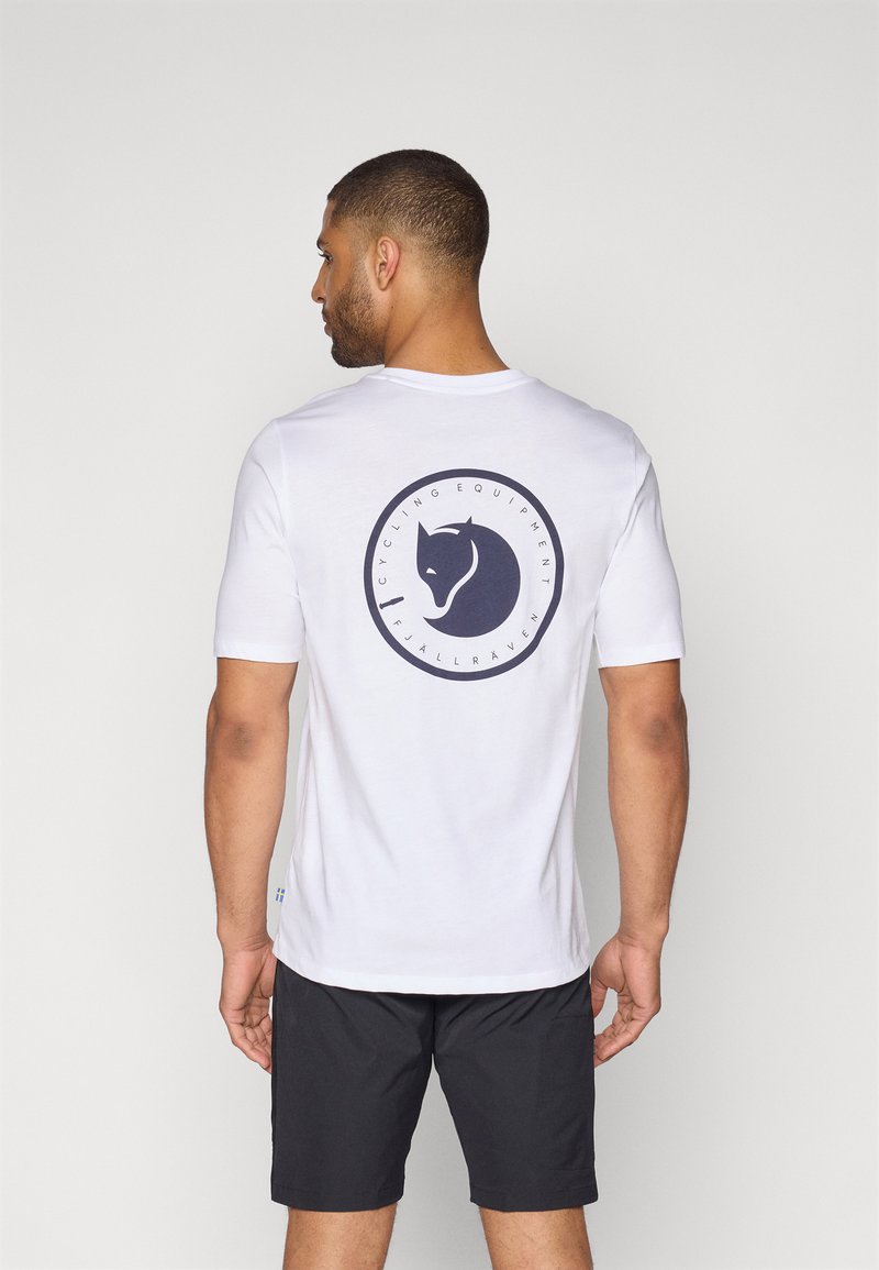Witte T-shirt met korte mouwen en een cirkelvormige marineblauwe grafische afbeelding met een gestileerd dierenhoofd en tekst, gedragen met zwarte shorts.