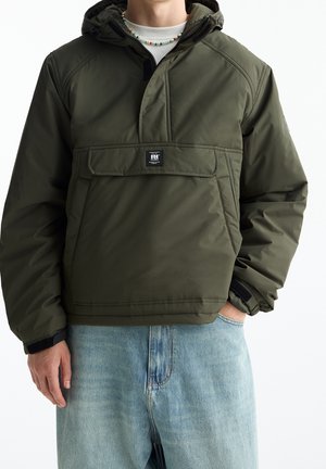 Homme portant une veste à capuche vert olive avec poche poitrine et un jean bleu clair, debout avec une main dans la poche.