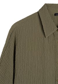 Koszulka w oliwkowym odcieniu z teksturą, z kołnierzykiem, wykonana z miękkiej, pogniecionej tkaniny oraz guzikiem z przodu.